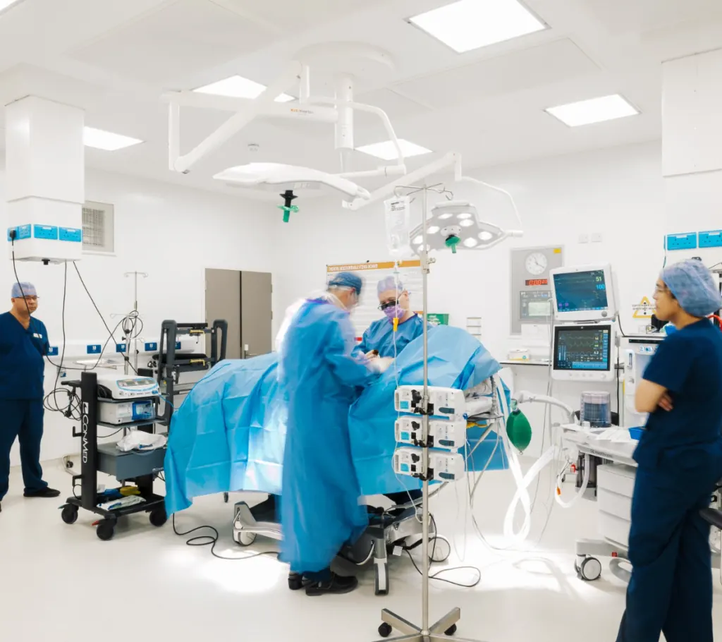Operating theatre at the Waterfront Private Hospital — surgical team gowned in blue around an active procedure, with overhead operating lights and patient monitors.